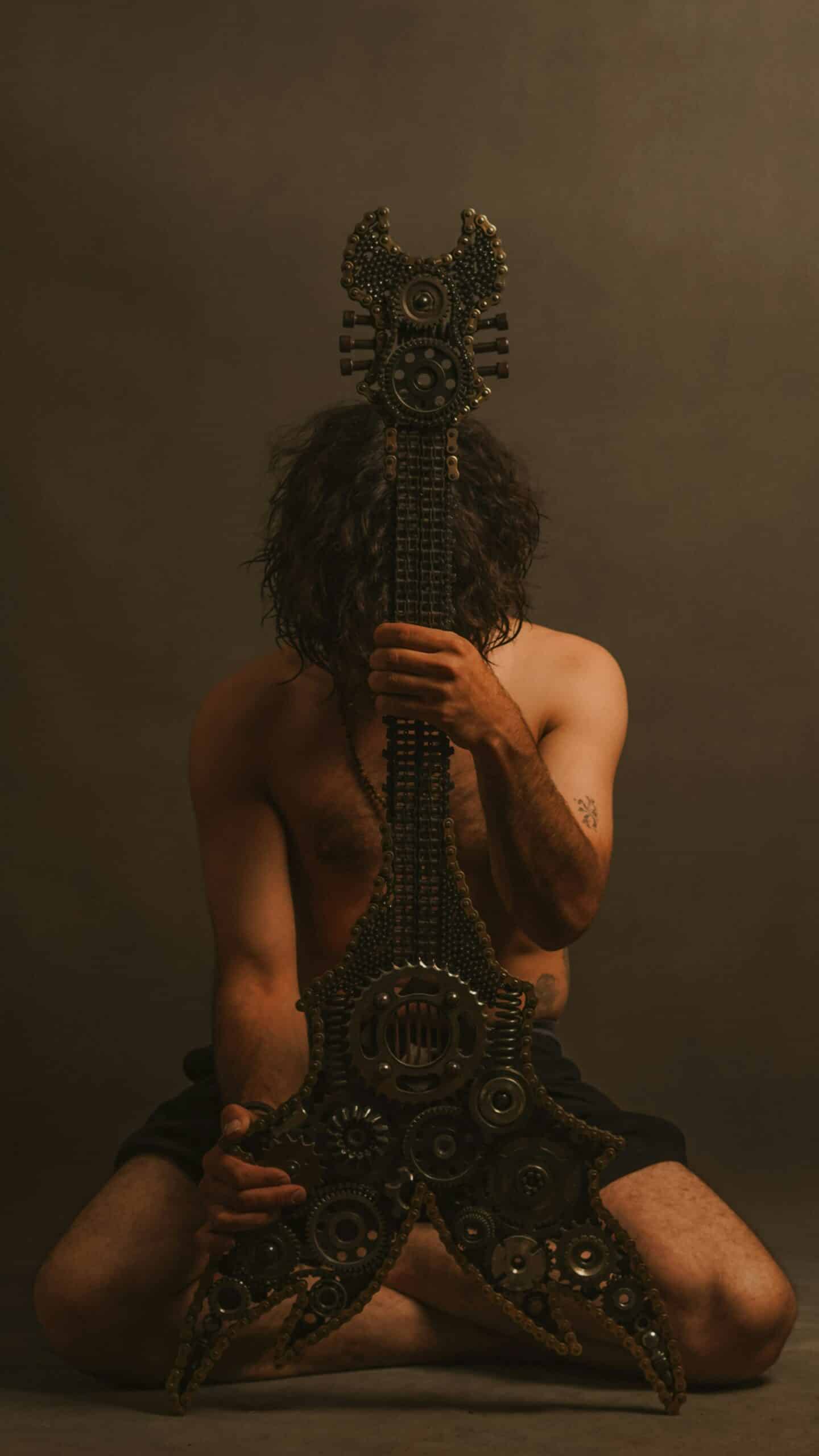 Portrait artistique inspiré de l’univers ISTOAR montrant un homme tenant une guitare métallique composée d’engrenages et de chaînes dans une ambiance sombre mêlant mythologie moderne et musique expérimentale.